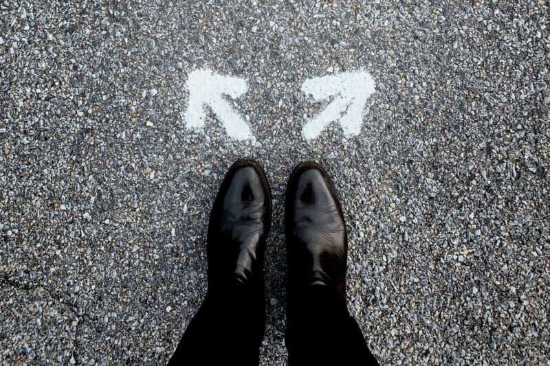 A pair of feet as seen from above. They are wearing black, leather formal shoes and black suit pants. In front of the shoes are two arrows in white spraypaint. One is pointing left and the other, right. 