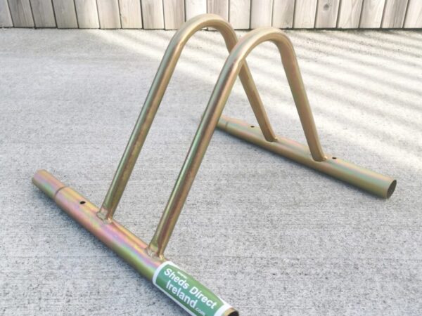 The Sheds Direct Ireland golden Bike Rack on a concrete ground and with a wooden wall behind it.