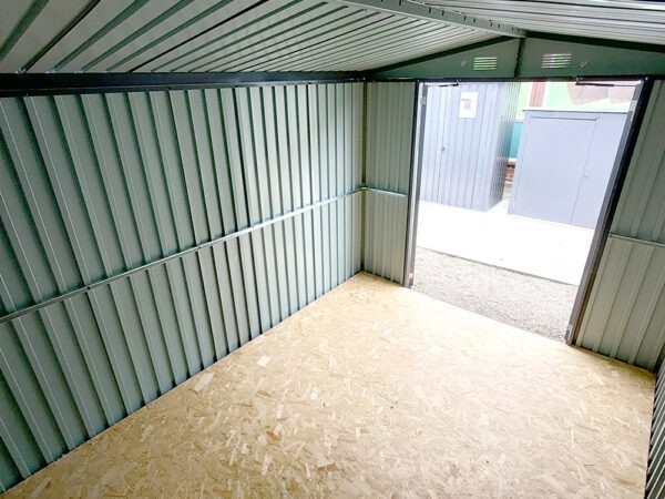 Inside the 8ft x 10ft steel premium garden shed. The view is from high in the top corner looking outwards to the door, which is on the right side of the frame. The walls are a light grey and they have a brace bar running along the centre