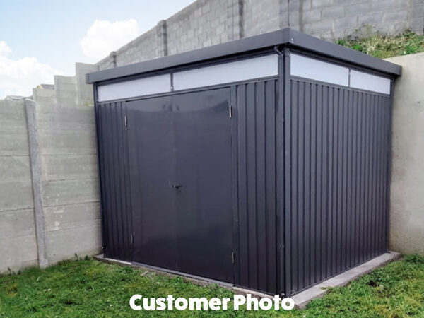 A customer's photo of the premium panoramic shed. It is up against a light-grey wall and dark green grass surrounds it