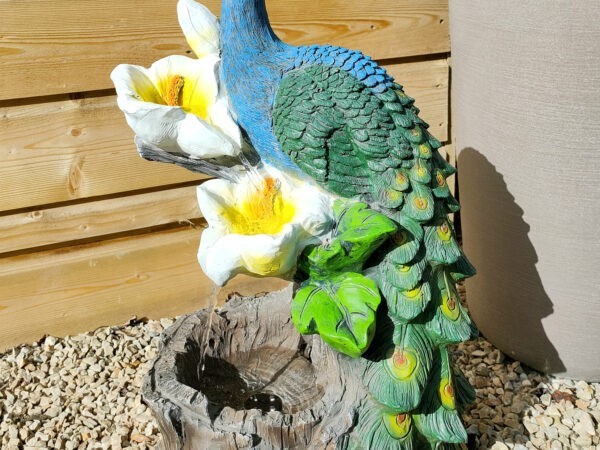 The Peacock Water feature against a green and grey plant pot. The peacock is blue and green and it sits on white and yellow lillies from which the water pours out