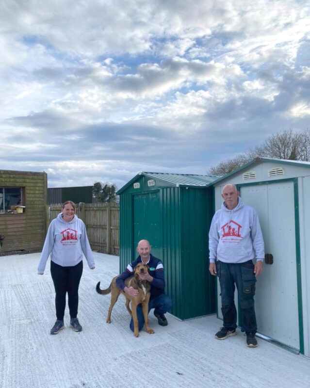 The team of Ramona and Chris from Coolronan Dog Rescue stand beside their new green and white sheds, while Alan from Sheds Direct Ireland hugs Hunter the dog. 