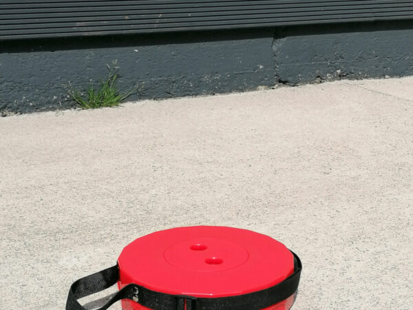 The red pop up stool in it's most compacted state. It is only 6cm tall and appears flat on a concrete ground below it. Behind there is a wooden fence.
