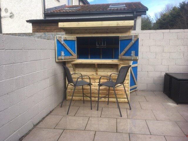 The Tolka Garden Bar from Sheds Direct Ireland in the corner of a garden. The windows are open showing the inside which is lined with blue material. There are two seats in front of the bar and a slate-grey wall behind it and to the left. There is a neighbours house visible behind it.