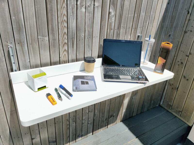 The Balcony Shelf filled with items including a laptop, a large book, a box of crisps, pens, a stationary holder, a coffee cup and pens.