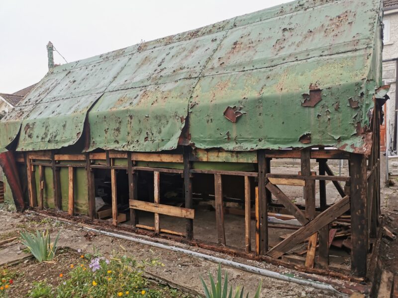 a battered old, green shed. The sheet metal on it is green and rested oragne and the supporting wooden frame is clearly visible through enormous holes in it