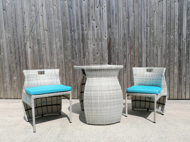 The balcony table and chairs set from Sheds Direct Ireland against a wooden wall. The chairs have the apperance of wicker and there is a bright baby blue cushion on each one. The table has a barrel-like appearance with a flat top. On the top of the unit sits a shiny, black glass tabletop.