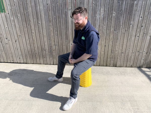 A man sitting on the yellow pop-up stool against a vertically planked wooden wall. The man is wearing a sheds direct ireland jumper, he has grey jeans and white shows on, a beard and clear-framed glasses. He is sitting in the centre of the frame looking to the left of the picture.