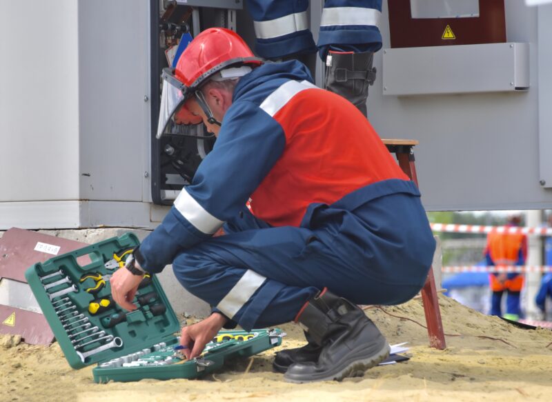 An electrician in a large red puffy jacket (who is not part of the steel shed assembly team), opening a box of electrical tools as he prepares to work