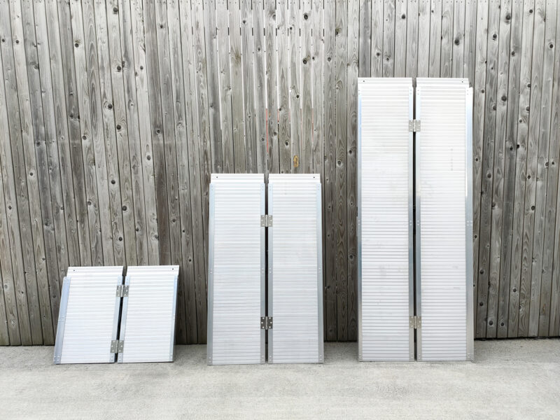 The three wheelchair ramps lined up against each other in ascending order, according to size. They are shiny aluminium and the size difference between the each is proportional. They are lined up against a wooden wall and standing on a concrete ground.