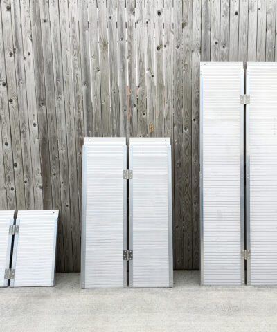 The three wheelchair ramps lined up against each other in ascending order, according to size. They are shiny aluminium and the size difference between the each is proportional. They are lined up against a wooden wall and standing on a concrete ground.