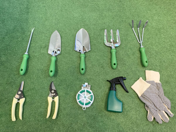 All the items from the garden tool bag with chair set laid out on a grassy surface. There is a long pronged fork, two trowels, two rakes, two clippers, a roll of twine, a spray-bottle and a set of gloves