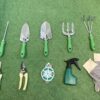 All the items from the garden tool bag with chair set laid out on a grassy surface. There is a long pronged fork, two trowels, two rakes, two clippers, a roll of twine, a spray-bottle and a set of gloves