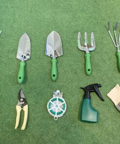 All the items from the garden tool bag with chair set laid out on a grassy surface. There is a long pronged fork, two trowels, two rakes, two clippers, a roll of twine, a spray-bottle and a set of gloves