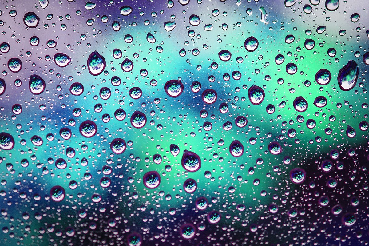 a teal, mint-green and purple blurred background sits behind water droplets on a pane of glass in the foreground. The droplets are coloured like the background, but in small, magnified portions. 