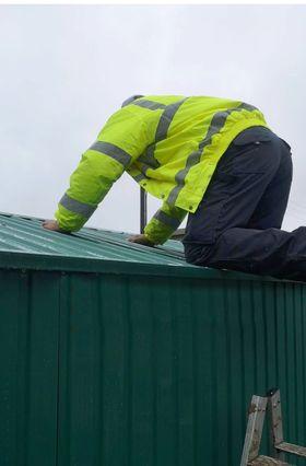 self assembly: A man in a hi-vis jacket kneeling on the edge of an assembled, green shed. He has both hands on the shed