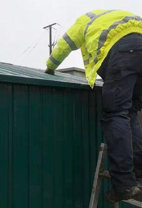 A man with a drill in his hand on a ladder assembling the roof of a steel garden shed