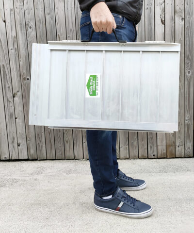 The Small Wheelchair Ramp being held by a man in a navy shirt, dark jeans and navy shoes. He is holding the ramp in one hand and it looks light in his arms.