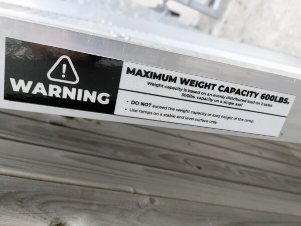 The warning sticker on the side of the ramp. It reads: MAXIMUM WEIGHT CAPACITY 600LBS. Weight capacity is based on an evenly distributed load on 2 axles. 300lbs capacity on a single axel. Do not exceed the weight capacity or load height of the ramp. Use ramps on a stable and level surface only'