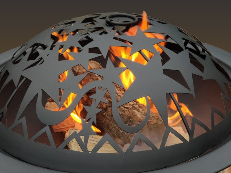 The Zodiac firepit in use in a back garden. Bright orange flames billow out the top, while inside dark red wooden embers glow dimly under the matte black covering