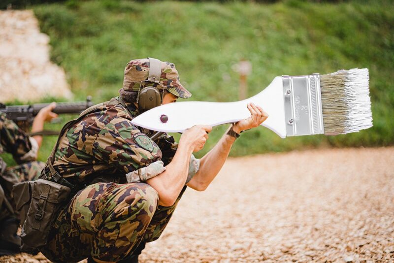 A man in military uniform holding a giant paint brush like you would hold a rifle. He is crouched, wearing ear-protectors, goggles and camouflaged clothes. The brush is white with white paint on the brown bristles. He's the soldier painting a steel shed