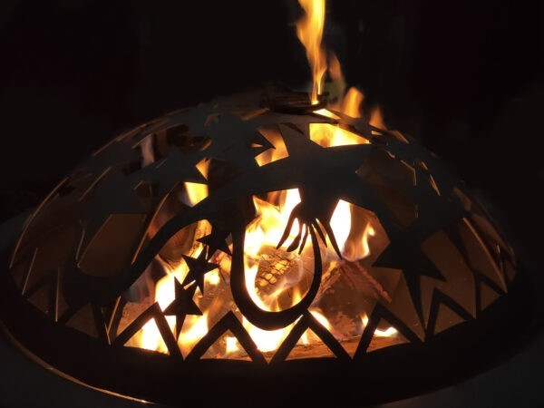 A very contrasty picture of the inferno fire pit in use. The flames are lapping out the top and they are orange and white. The rest of the photo is a heavily shadowed black, almost a silhouette from the flames