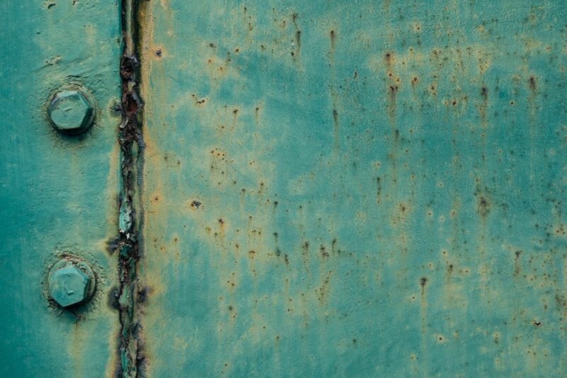 A green and blue faded piece of metal seen up close. There are two rivets to the side which have a slight brown rust around them. There is a hinge to the left which is a darker colour also