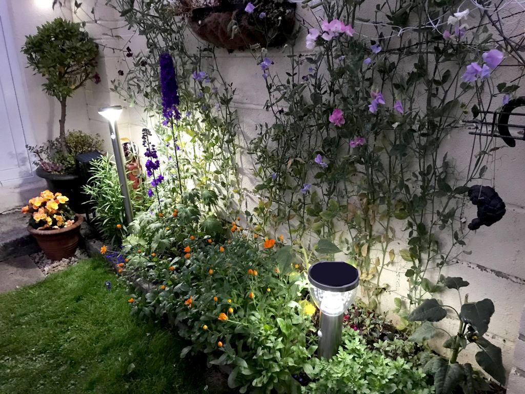 Two LED Garden Lights in a flower bed. The one closer to the camera is small, the other is very tall. They are both operational and they are illuminating the orange and purple flowers below them. There are hanging plants above the lights also, but these are in a half-light.