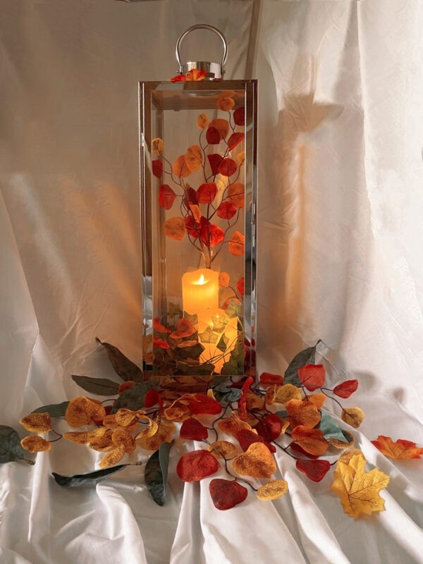 An Autumnal scene in a candle holder. The candle holder is infront of a white cloth background, there are various types of leaves at the base in various autumn colours and there is an orange candle inside the candle holder. 