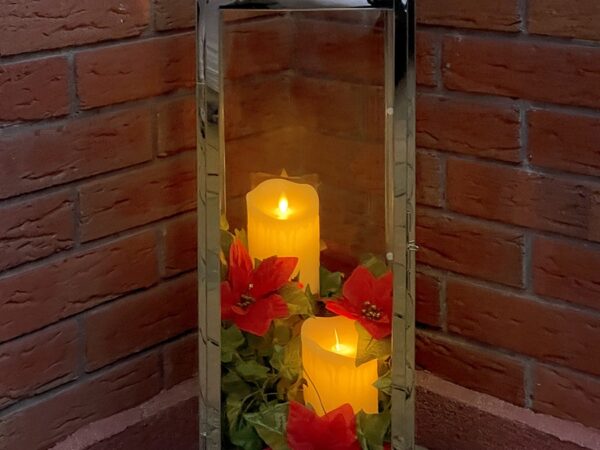 A Candle Holder with red flowers and yellow candles inside. The candle holder is in a corner, with two red-brick walls either side of it. The candles are glowing a warm orange in colour.