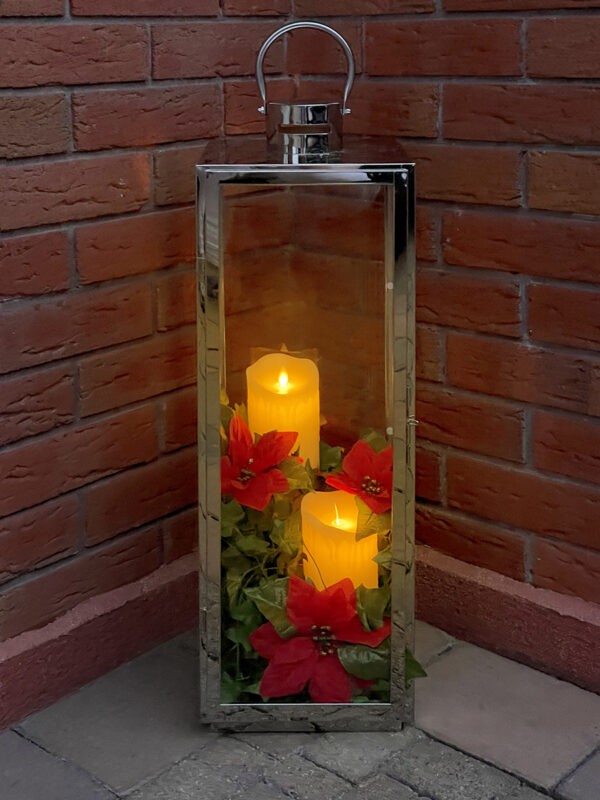 A Candle Holder with red flowers and yellow candles inside. The candle holder is in a corner, with two red-brick walls either side of it. The candles are glowing a warm orange in colour.