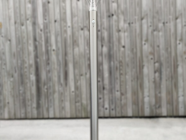 The Tall Silver Garden Light bolted down to concrete against a brown, wooden wall