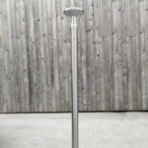 The Tall Silver Garden Light bolted down to concrete against a brown, wooden wall