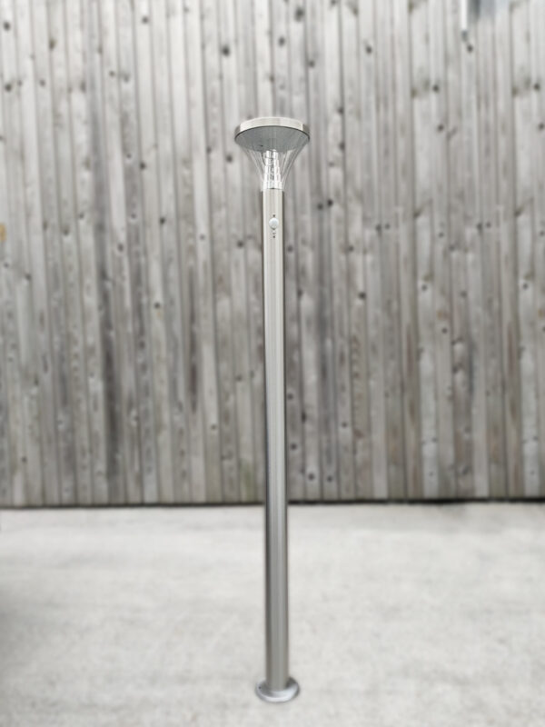The Tall Silver Garden Light bolted down to concrete against a brown, wooden wall