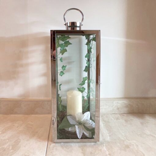 A silver, lantern-style candle holder with green leaves trailing down the inside on both edges, with a cream-coloured candle in the middle. There is a large white lily opened at the base of the candle. The candle stands on a white marble floor against a white wall.