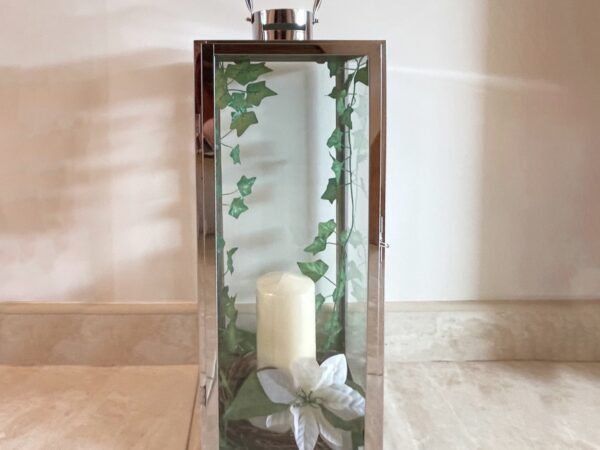 A silver, lantern-style candle holder with green leaves trailing down the inside on both edges, with a cream-coloured candle in the middle. There is a large white lily opened at the base of the candle. The candle stands on a white marble floor against a white wall.