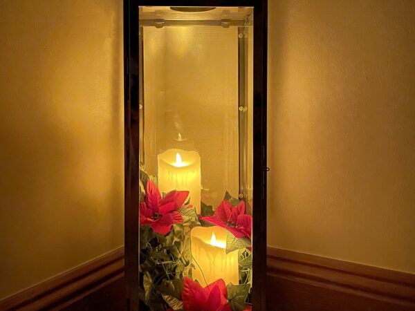 A candle holder lit exclusively by the candles inside, in an otherwise dark room. The candles are a yellow colour and there are red flowers along the bottom of it.