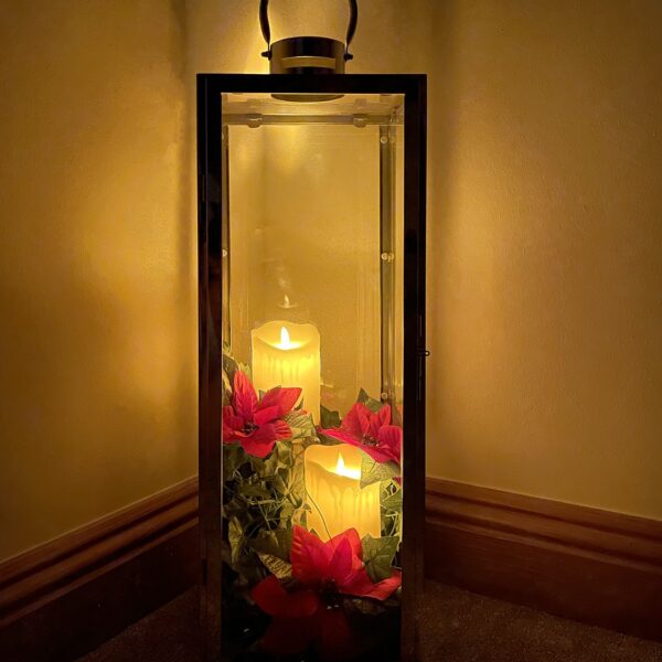 A candle holder lit exclusively by the candles inside, in an otherwise dark room. The candles are a yellow colour and there are red flowers along the bottom of it.