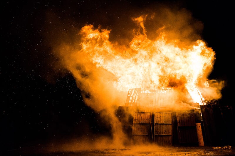 A wooden shed that is engulfed in bright orange flames. There is no other light in the scene, so it is black at the background and the shed is glowing orange