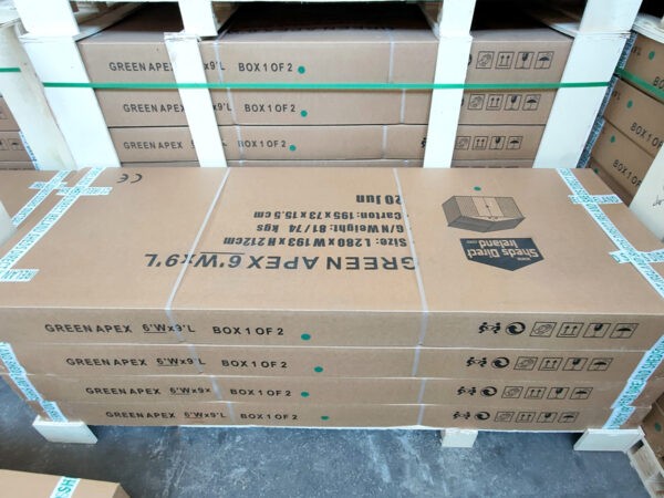 A stack of flat-packed boxes of sheds ontop of each other. There are 4 large and wide boxes on top of each other. They are brown and the text on top is upside-down and it reads 'GREEN APEX 6 FOOT X 9 FOOT'