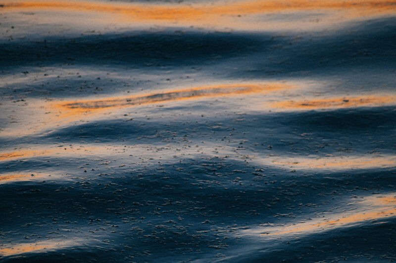 A close up image of a dirty liquid with large particles of dirt and grit visible on the surface. The liquid is a dark blue and it is backlit by orange light