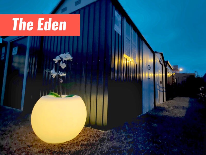 The Eden Flower pot outside a cottage garden shed. It's glowing a gentle orange hue. The sky and sheds behind are a dark, blue, pre-dawn colour. 