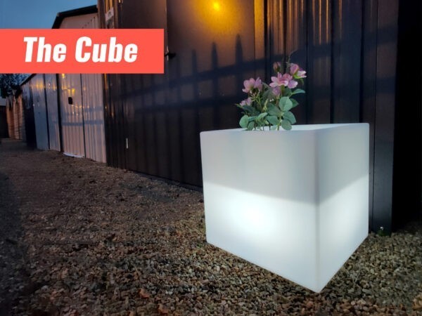 The Cube LED plant pot outside a shed, just before dawn. The plant pot is glowing white and the sheds behind it are dark, except for the yellow lamp-lights.