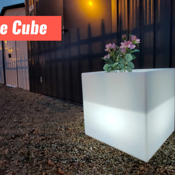 The Cube LED plant pot outside a shed, just before dawn. The plant pot is glowing white and the sheds behind it are dark, except for the yellow lamp-lights.