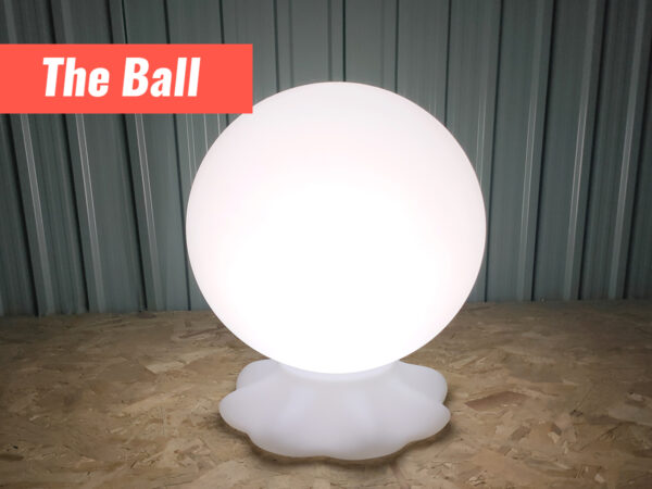 A ball shaped LED light standing on a white faux-rock base. Its on a plywood foundation and against a metal wall