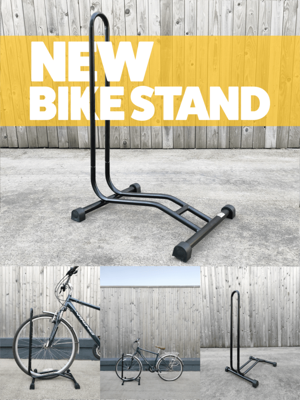 A black bike stand against a great wall and concrete flooring. There is a yellow banner that goes through the hoop of the bike stand with text on it that reads 'bike stand'