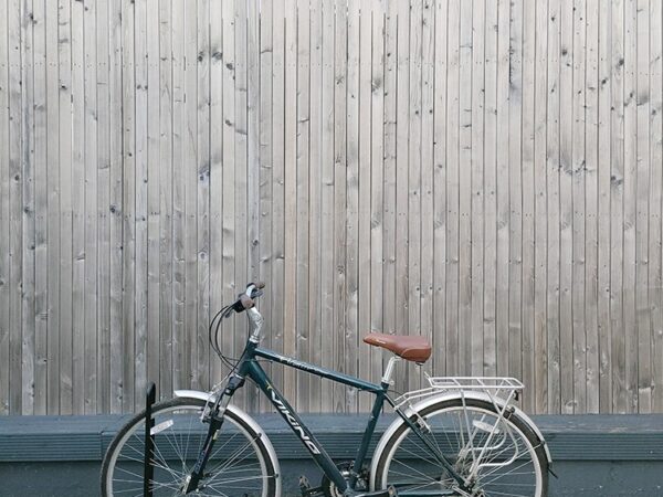 A black, old-fashioned bike parked into a bike stand. The bike is standing upright and it is close to a wooden wall behind it