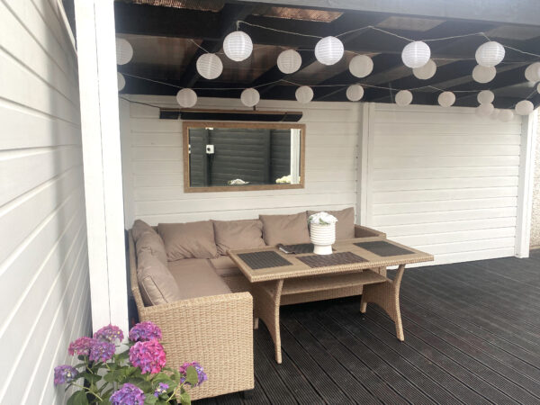 The Ronan Heating heater with speaker mounted on the wall of a pergola. The walls are white, the floor is black and there is bunting-lights hanging from the room. Above a mirror on the wall, the ronan heating is visible
