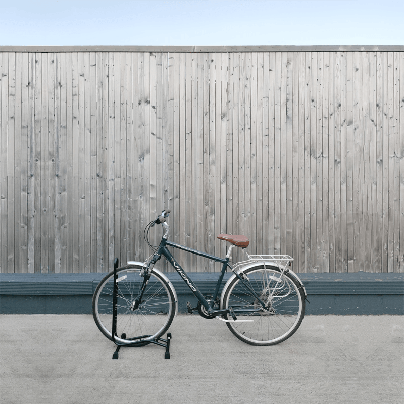 A black, old-fashioned bike parked into a bike stand. The bike is standing upright and it is close to a wooden wall behind it