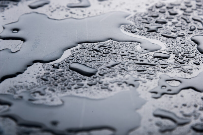 Water pooling on a black, metallic surface. There are two large globules of water and other smaller droplets all around them